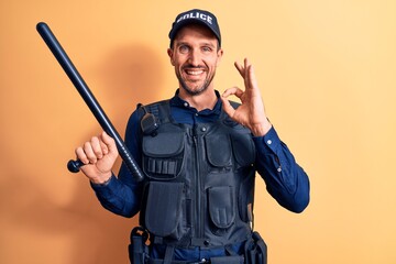 Handsome policeman wearing uniform and bulletprof holding baton over yellow background doing ok sign with fingers, smiling friendly gesturing excellent symbol