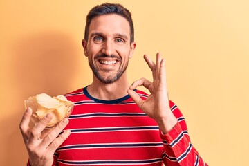 Young handsome man holding bowl of potato chips standing over isolated yellow background doing ok...