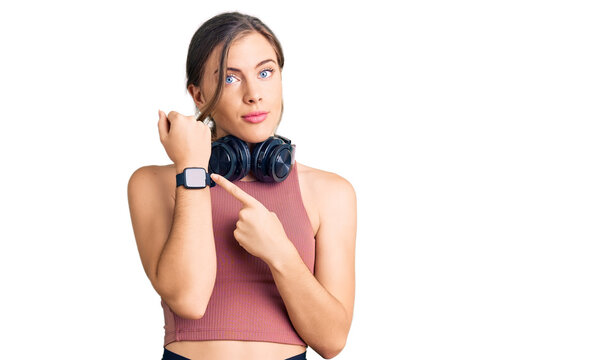 Beautiful caucasian young woman wearing gym clothes and using headphones in hurry pointing to watch time, impatience, looking at the camera with relaxed expression