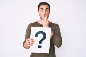 Young handsome man holding question mark covering mouth with hand, shocked and afraid for mistake. surprised expression