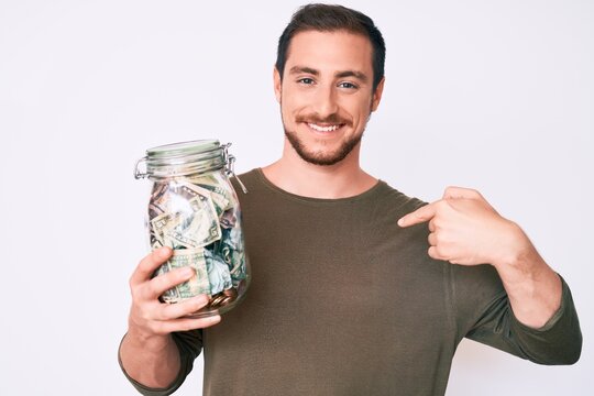 Young handsome man holding jar with savings pointing finger to one self smiling happy and proud