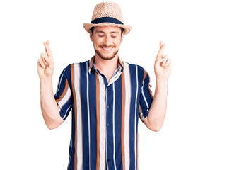 Young handsome caucasian man wearing summer hat gesturing finger crossed smiling with hope and eyes closed. luck and superstitious concept.
