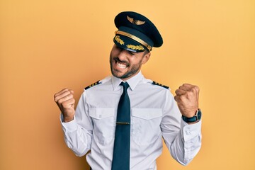 Handsome hispanic man wearing airplane pilot uniform very happy and excited doing winner gesture with arms raised, smiling and screaming for success. celebration concept.