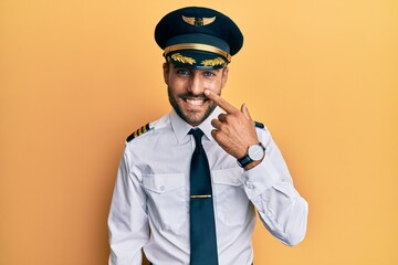 Handsome hispanic man wearing airplane pilot uniform pointing with hand finger to face and nose, smiling cheerful. beauty concept