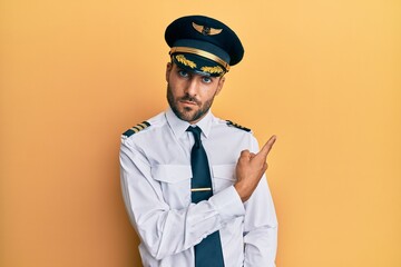 Handsome hispanic man wearing airplane pilot uniform pointing with hand finger to the side showing advertisement, serious and calm face