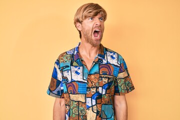 Young blond man wearing summer shirt angry and mad screaming frustrated and furious, shouting with...
