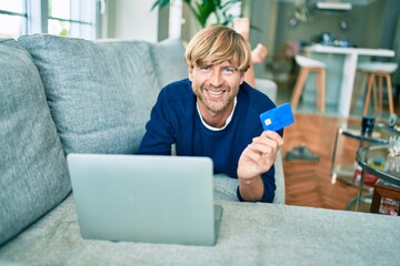 Middle age handsome caucasian man relaxing at home doing online shopping using credit card on computer laptop