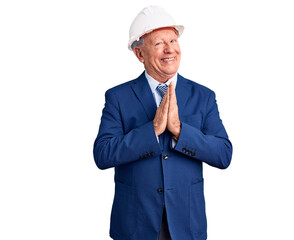 Senior handsome grey-haired man wearing suit and architect hardhat praying with hands together asking for forgiveness smiling confident.