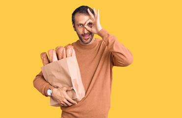 Young handsome man holding paper bag with bread smiling happy doing ok sign with hand on eye looking through fingers