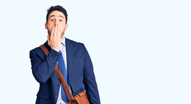 Young hispanic man wearing suit and leather bag covering mouth with hand, shocked and afraid for mistake. surprised expression