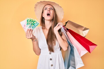 Young blonde girl holding shopping bags and israeli shekels angry and mad screaming frustrated and furious, shouting with anger looking up.