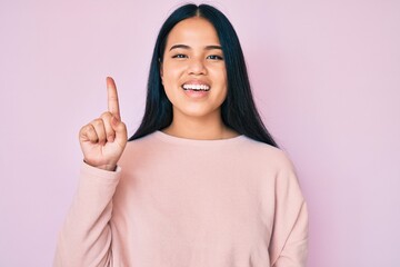 Young beautiful asian girl wearing casual pink sweater smiling with an idea or question pointing finger up with happy face, number one