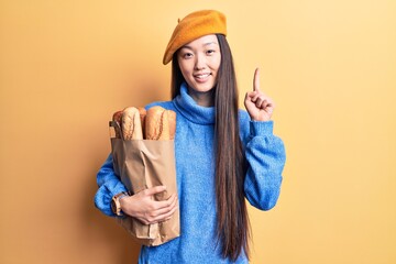 Young beautiful chinese woman wearing french beret holding paper bag with bread smiling with an idea or question pointing finger with happy face, number one