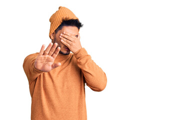 Handsome latin american young man wearing winter sweater and wool hat covering eyes with hands and...