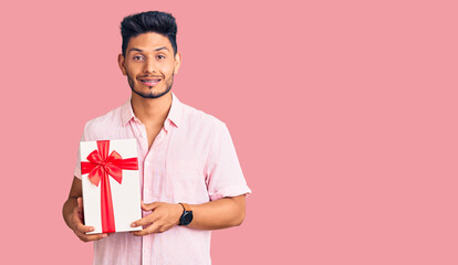 Handsome latin american young man holding gift looking positive and happy standing and smiling with a confident smile showing teeth