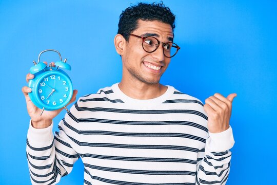 Young handsome hispanic man holding alarm clock pointing thumb up to the side smiling happy with open mouth