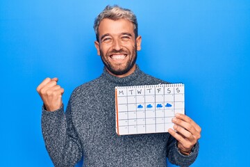 Young handsome blond man with beard holding weather calendar showing rainy week screaming proud, celebrating victory and success very excited with raised arm