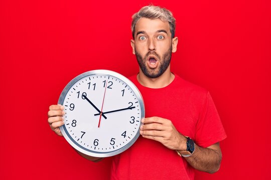 Young handsome blond man with beard doing countdown using big clock over red background scared and amazed with open mouth for surprise, disbelief face