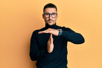Handsome man with tattoos wearing turtleneck sweater and glasses doing time out gesture with hands, frustrated and serious face