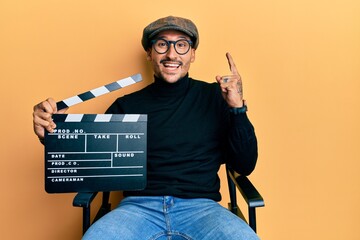 Handsome man with tattoos holding video film clapboard sitting on director chair smiling with an...