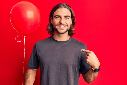 Young handsome man holding balloon pointing finger to one self smiling happy and proud