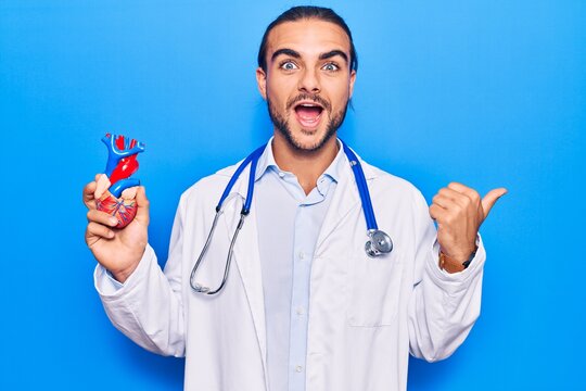 Young handsome man wearing doctor stethoscope holding heart pointing thumb up to the side smiling happy with open mouth