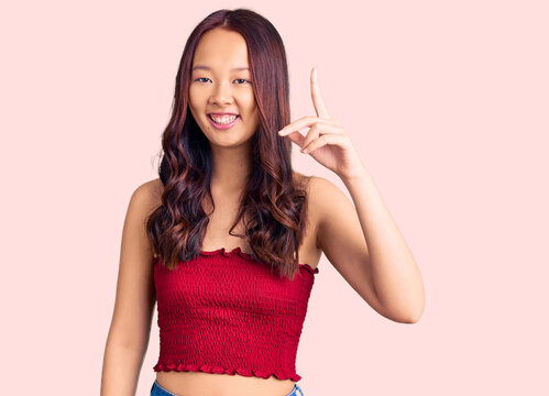Young beautiful chinese girl wearing casual clothes pointing finger up with successful idea. exited and happy. number one.