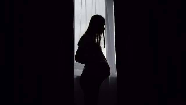 Silhouette of young pregnant caucasian woman standing near window with curtains in house. Side view of happy future mothers looking and gently stroking her stomach. Concept motherhood