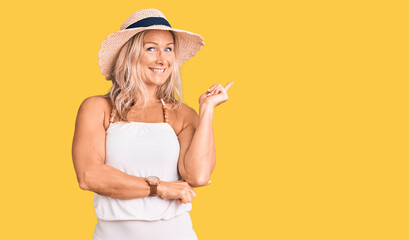 Middle age fit blonde woman wearing summer hat with a big smile on face, pointing with hand and finger to the side looking at the camera.