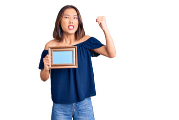 Young beautiful chinese girl holding empty frame annoyed and frustrated shouting with anger, yelling crazy with anger and hand raised