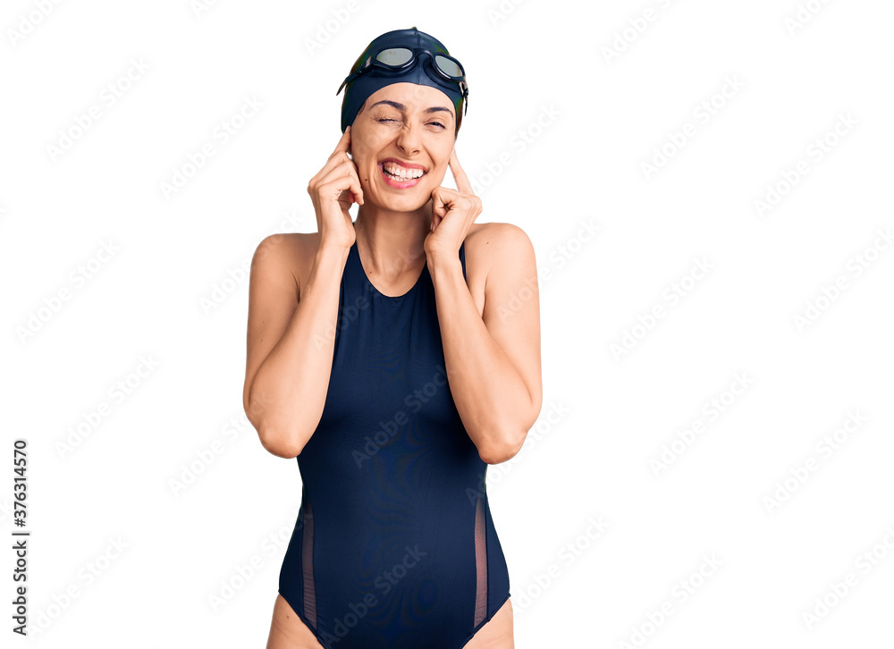 Wall mural young beautiful hispanic woman wearing swimwear and swimmer glasses covering ears with fingers with 