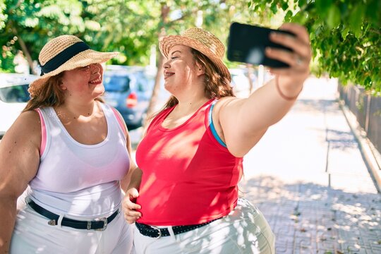 Two plus size overweight sisters twins women smiling taking a selfie picture with the phone outdoors on a sunny day