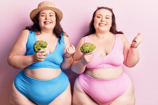 Young plus size twins wearing bikini holding broccoli screaming proud, celebrating victory and success very excited with raised arm