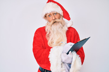 Old senior man with grey hair and long beard wearing santa claus costume writting list in shock face, looking skeptical and sarcastic, surprised with open mouth