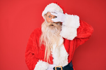 Old senior man with grey hair and long beard wearing traditional santa claus costume peeking in shock covering face and eyes with hand, looking through fingers with embarrassed expression.