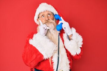 Old senior man wearing santa claus costume using vintage telephone serious face thinking about question with hand on chin, thoughtful about confusing idea