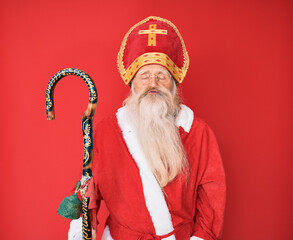 Old senior man with grey hair and long beard wearing traditional saint nicholas costume looking at the camera blowing a kiss on air being lovely and sexy. love expression.