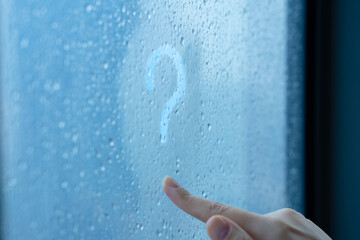 Female hand draws a question mark on a foggy window during the rain. A glass in drops of water.