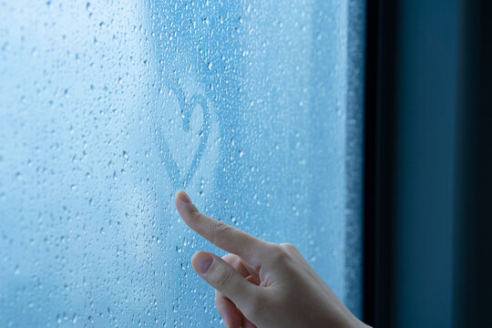 Female hand drawings a heart on a foggy window during the rain. Glass in drops of water. The concept of romance.