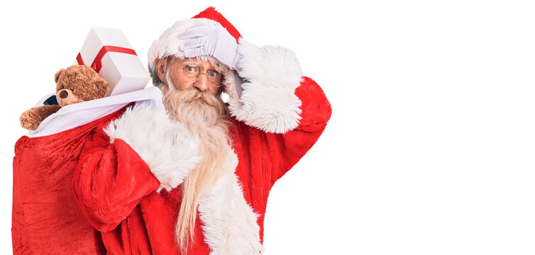 Old senior man with grey hair and long beard wearing santa claus costume holding bag with presents stressed and frustrated with hand on head, surprised and angry face