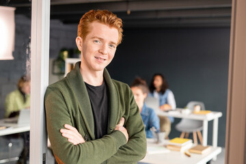 Young cross-armed businessman in casualwear standing by open door of office