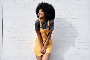 Happy stylish African American young woman wears yellow trendy sundress with Afro hair laughing looking away standing against white brick wall outdoor background. Smiling black hipster woman portrait - obrazy, fototapety, plakaty