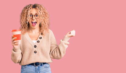 Young blonde woman with curly hair wearing glasses and drinking a cup of coffee screaming proud, celebrating victory and success very excited with raised arms