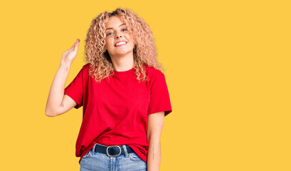 Young blonde woman with curly hair wearing casual red tshirt waiving saying hello happy and smiling, friendly welcome gesture