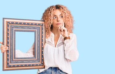 Young blonde woman with curly hair holding empty frame serious face thinking about question with...