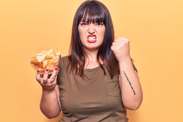 Young plus size woman holding potato chip annoyed and frustrated shouting with anger, yelling crazy with anger and hand raised
