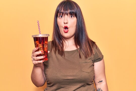Young plus size woman drinking glass of cola beverage scared and amazed with open mouth for surprise, disbelief face