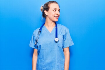 Young blonde woman wearing doctor uniform and stethoscope looking away to side with smile on face,...