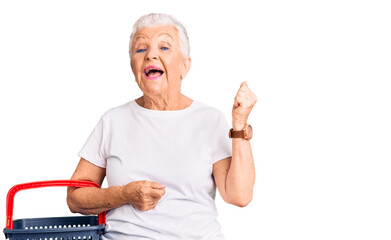 Senior beautiful woman with blue eyes and grey hair holding supermarket shopping basket screaming proud, celebrating victory and success very excited with raised arms