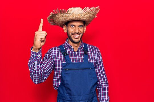 Young latin man wearing farmer hat and apron pointing finger up with successful idea. exited and happy. number one.